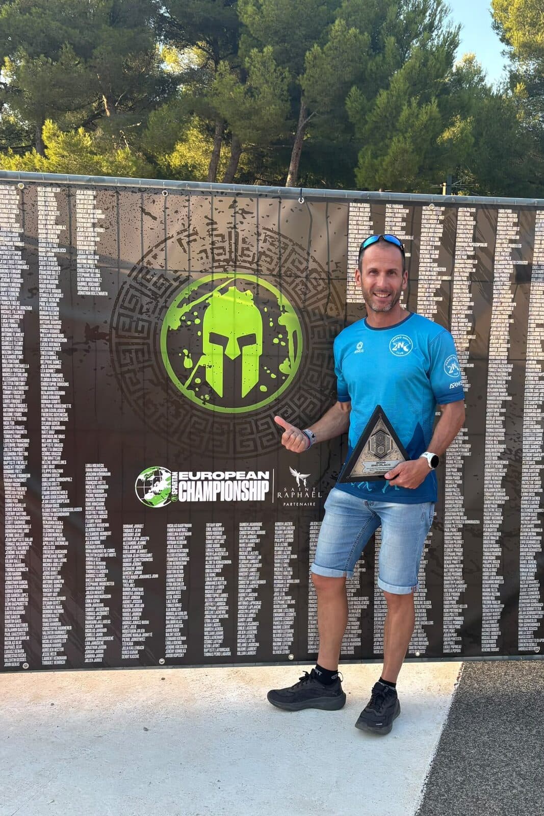 Jérôme, maillot du club, tient son trophée devant le panneau du championnat d'Europe Spartan Race
